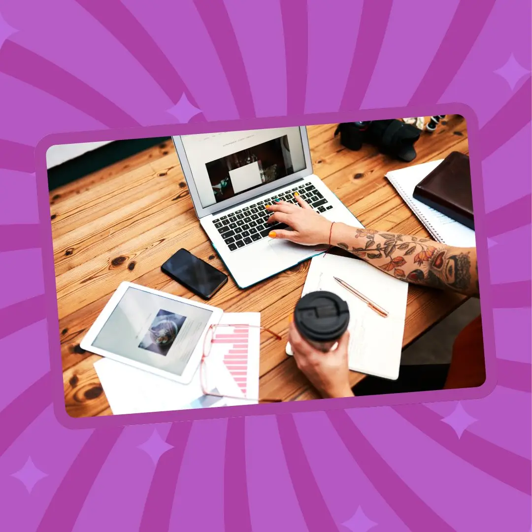 Overhead view of a person with tattooed arms using a laptop on a wooden table, surrounded by notes, a tablet, and a coffee cup.